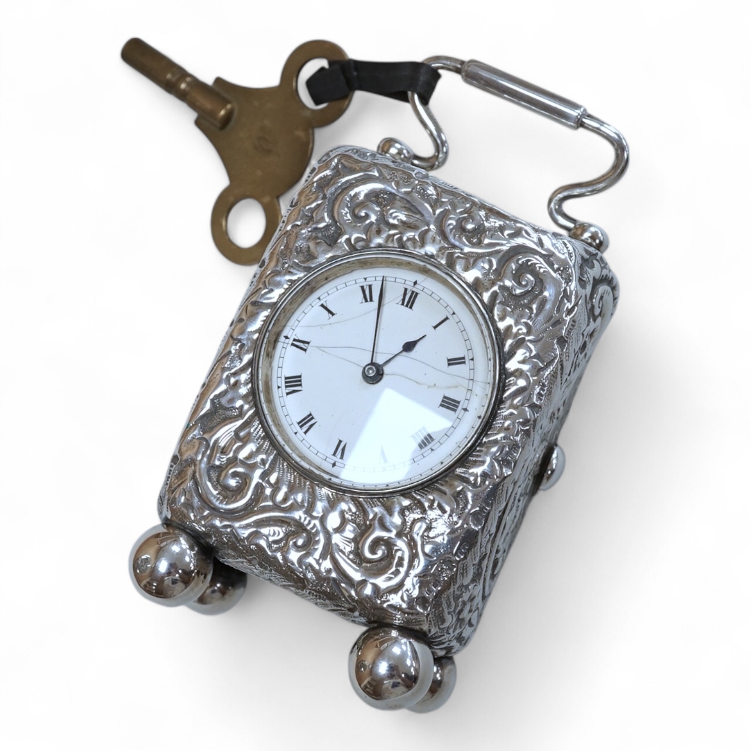 An early 20th century white metal (tests as silver) carriage timepiece, with white Roman dial (cracks) and case embossed with foliage, on four ball feet, 76mm excluding handle, with key.